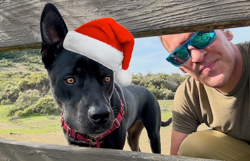 Shay Elkin, wearing blue sunglasses and an olive colored shirt, poses outdoor, next to a big black dog wearing a Santa Hat.